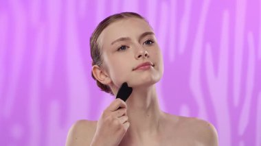 Genç bir kadın, mor bir arkaplana dayanan bir yüz taşı aleti kullanarak cilt rutinini gösteriyor..