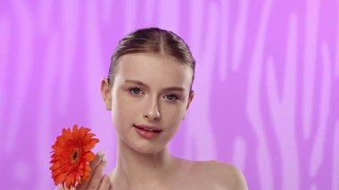 Genç bir kadın parlak turuncu bir gerbera papatyasını gözünün önünde canlı mor bir arka planda tutuyor..