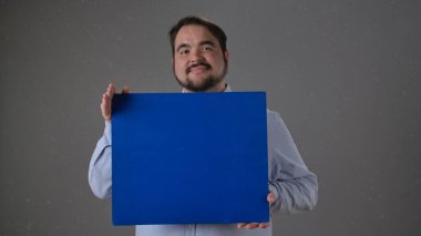 Elinde parlak mavi bir tabela tutarken gülümseyen bir adam. İki eli de gri bir arka planın önünde..