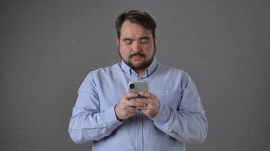 Akıllı telefonunu konsantrasyonla kullanan bir adam ayakta duruyor. Açık mavi bir gömlek giyiyor ve düşünceli görünüyor..