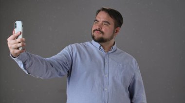 Mavi gömlekli bir adam akıllı telefonunu kullanarak özgüvenli bir selfie çeker, pozitiflik ve güvenini ifade eder..