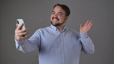 Mavi tişörtlü bir adam gülümseyip el sallarken akıllı telefonuyla videoyu arıyor..