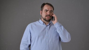 Açık mavi düğmeli gömlekli bir adam gri arka planda akıllı bir telefonla konuşuyor, düşünceli görünüyor..