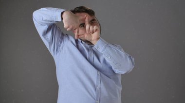 Mavi gömlekli bir adam, düz bir arkaplan karşısında eşsiz bir perspektif yakalayan, elleriyle bir görüntüyü yaratıcı bir şekilde çerçeveler..