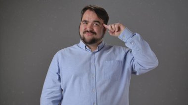 Mavi gömlekli bir adam, özgüvenli bir ifadeyle başını işaret eder, düz bir arkaplan karşısında durur..