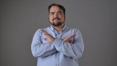 Açık mavi gömlekli bir adam kollarını gri bir arkaplan önünde çapraz yapar, elleri olan bir X sembolü yapar..