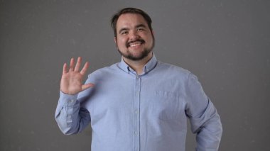 Açık mavi tişörtlü bir adam kameraya sıcak bir şekilde gülümsüyor ve gri arka plana doğru el sallıyor..