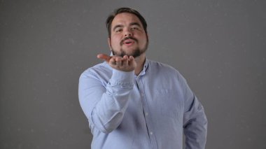 Açık mavi gömlekli bir adam, tarafsız gri bir arkaplana karşı dostça ve nazik bir ifadeyle öpücük yolluyor..