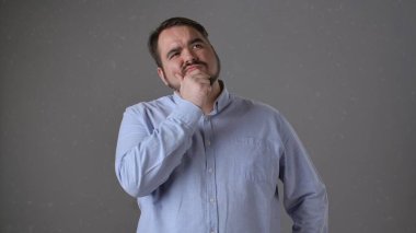 Açık mavi gömlekli bir adam, gri bir arka planda çenesini tutarken derin derin düşünürken görülüyor..