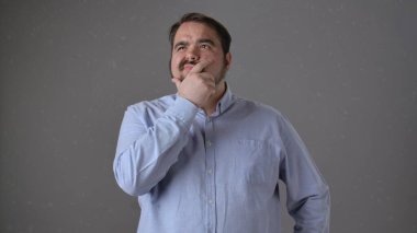 Açık mavi gömlekli bir adam, gri bir arka planda çenesini tutarken derin derin düşünürken görülüyor..