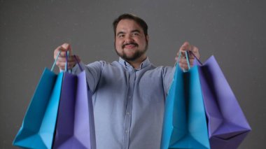 Gülümseyen bir adam renkli alışveriş torbalarını dostça sunar. Görüntü neşe ve memnuniyet verir..