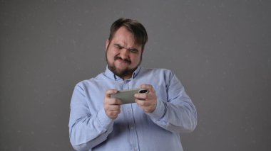 Açık mavi tişörtlü bir adam iki eliyle bir akıllı telefon tutuyor, mobil oyun oynarken hayal kırıklığı yaşıyor..