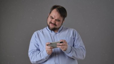 Açık mavi tişörtlü bir adam iki eliyle bir akıllı telefon tutuyor, mobil oyun oynarken hayal kırıklığı yaşıyor..