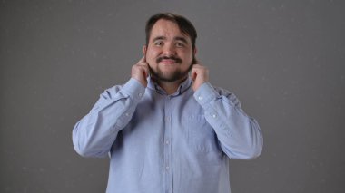 Mavi tişörtlü bir adam parmaklarıyla kulaklarını tıkar, hiçbir şey duymadığı jestini yapar, düz gri bir arka plana karşı..