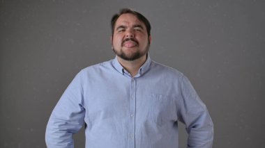 Açık mavi gömlekli bir adam, esprili bir şekilde dilini gri bir arka plana doğru uzatıyor..