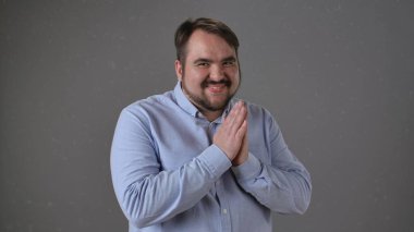 Açık mavi tişört giyen bir adam ellerini sıkarak muzip bir gülümseme sergiliyor..