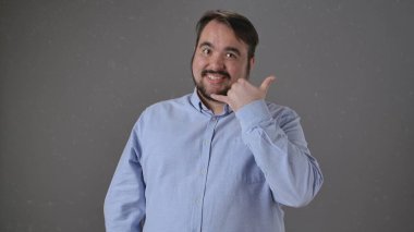 Mavi gömlekli, gülümseyen bir adam, baş parmağı ve serçe parmağıyla bir telefon hareketi yapıyor..