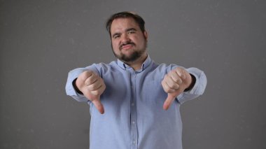 Mavi gömlekli bir adam, tarafsız bir arkaplana karşı baş parmak işareti yaparak onaylamadığını gösterir..