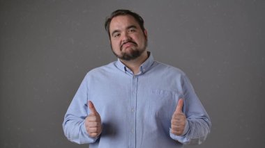 Açık mavi tişörtlü sakallı bir adam, tarafsız bir arkaplana karşı duran, kendine güvenen bir ifadeyle çifte onay verir..
