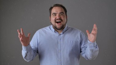 Açık mavi gömlekli bir adam heyecanını açık bir ağızla gösterir ve gri bir arka plana karşı ellerini kaldırır..