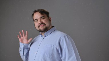 Açık mavi tişörtlü bir adam kameraya sıcak bir şekilde gülümsüyor ve gri arka plana doğru el sallıyor..