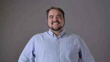 Açık mavi gömlekli bir adam, basit bir gri arka plana gülümsüyor, sıcaklığı ve dostluğu iletiyor. Yavaş çekim