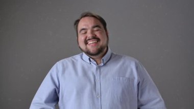 Açık mavi gömlekli bir adam gülüyor ve sonra gülerek düz gri bir arka plana, sıcaklığı ve dostluğu iletiyor. Ağır çekim Pozitif Duygular. Yavaş çekim