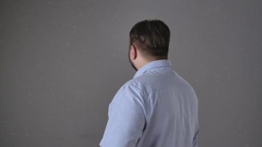 Açık mavi gömlekli bir adam kameraya döner, sıcak bir şekilde gülümser ve kameraya gri bir arka plana doğru el sallar. Yavaş çekim