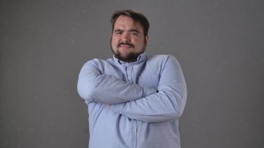 Açık mavi gömlekli, çapraz kollu bir adam güvenle ayakta duruyor. Yavaş çekim. İfadesi düşünceli, odaklanmış ve tarafsız bir arkaplana karşı kararlılık gösteriyor. Yavaş çekim