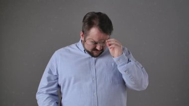 Mavi gömlekli sakallı bir adam hayal kırıklığı içinde gözlerini ovalıyor. Yorgunluk ve stresi simgeliyor. Yavaş çekim