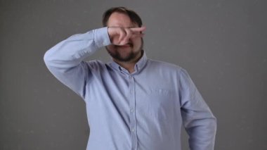 Açık mavi gömlekli adam neşeyle dans ediyor ve el hareketi yapıyor. Ağır çekimde sakallı bir adam tarafsız gri bir arka plana karşı duruyor. Yavaş çekim