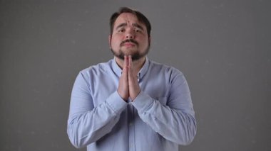 Açık mavi gömlekli bir adam umut dolu ve yalvaran bir ifadeyi gri arka plana yapıştırılmış bir şekilde gösterir. Yavaş çekim