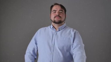 Bir adam gri arka planda duruyor ve el hareketini gösteriyor. Buraya gelip birini arıyor. Ağır çekimde mavi gömlekli, sakallı, orta yaşlı bir adam. Yavaş çekim