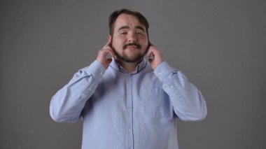 Mavi gömlekli bir adam kulaklarını parmaklarıyla tıkar, hiçbir şey duymadığını belirten bir jest yapar, düz gri bir arka plana karşı. Yavaş çekim