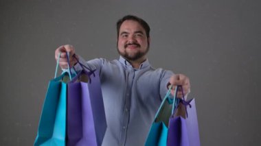 Gülümseyen bir adam renkli alışveriş torbalarını dostça sunar. Yavaş çekim neşe ve memnuniyeti iletir. Yavaş çekim