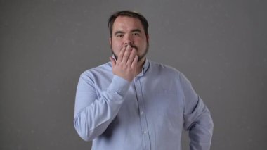 Açık mavi gömlekli bir adam, tarafsız gri bir arkaplana karşı dostça ve nazik bir ifadeyle öpücük yolluyor..