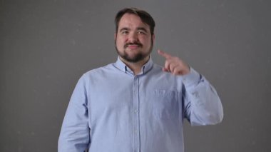 Mavi gömlekli bir adam, özgüvenli bir ifadeyle başını işaret eder, düz bir arkaplan karşısında durur..