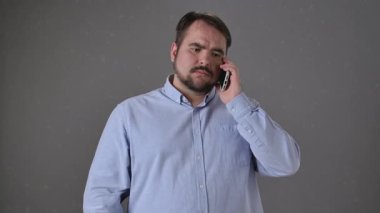 Açık mavi düğmeli gömlekli bir adam gri arka planda akıllı bir telefonla konuşuyor, düşünceli görünüyor..