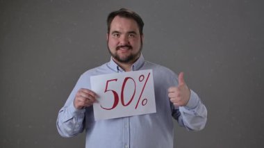 Mavi gömlekli bir adam% 50 yazılı bir tabela tutar. Onayını ya da memnuniyetini gösteren bir jest yapar..