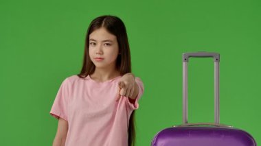 Pembe gömlekli genç kız mor bavulun yanında duruyor, kamerayı işaret ediyor ve yeşil ekran yeşil ekran stüdyo arka plan modelinde izole edilmemiş jest yapıyor. Yavaş çekim