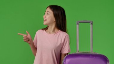Pembe tişörtlü genç kız mor bavulun yanında duruyor ve parmağıyla bize işaret ediyor, kopyalama alanı. Krom anahtar yeşil ekran yeşil ekran stüdyo arka plan modelinde izole edilmiş. Yavaş çekim