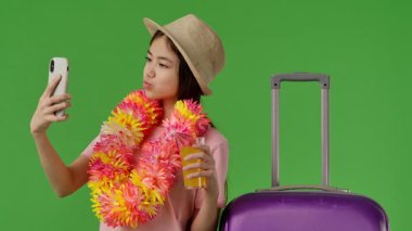Çiçek kolyesi takan ve elinde portakal suyu olan bir kadın valiziyle selfie çeker. Tatil macerasına hazırdır. Krom anahtar yeşil ekran yeşil ekran stüdyo arka plan modelinde izole edilmiş. Yavaş çekim