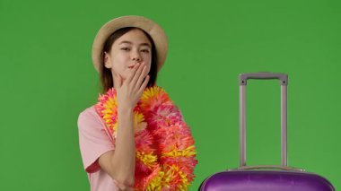 Çiçek kolyeli genç kız mor bavulun yanında duruyor ve hava öpücüğü gönderiyor. Krom anahtar yeşil ekran yeşil ekran stüdyo arka plan modelinde izole edilmiş. Yavaş çekim