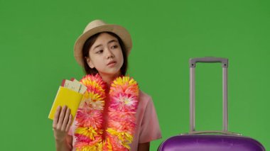 Çiçekli genç kız mor bavulun yanında duruyor ve elinde uçak biletleriyle pasaport tutmaktan yorulmuş. Krom anahtar yeşil ekran yeşil ekran stüdyo arka plan modelinde izole edilmiş. Yavaş çekim