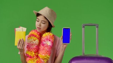 Çiçekli çiçek kolyeli genç bir kız mor bavulun yanında duruyor, elinde pasaportu ve akıllı telefonu var. Krom anahtar yeşil ekran yeşil ekran stüdyo arka plan modelinde izole edilmiş. Yavaş çekim