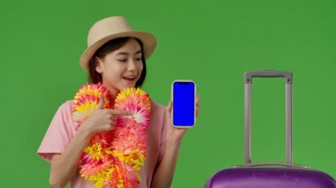 Mor bavulun yanında duran çiçek yapraklı genç kız mavi ekranlı akıllı telefonu işaret ediyor. Krom anahtar yeşil ekran yeşil ekran stüdyo arka plan modelinde izole edilmiş. Yavaş çekim