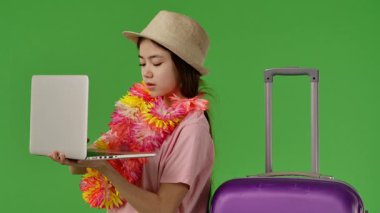 Çiçek kolyeli genç kız mor bavulun yanında duruyor ve dizüstü bilgisayarında daktilo yazıyor. Krom anahtar yeşil ekran yeşil ekran stüdyo arka plan modelinde izole edilmiş. Yavaş çekim