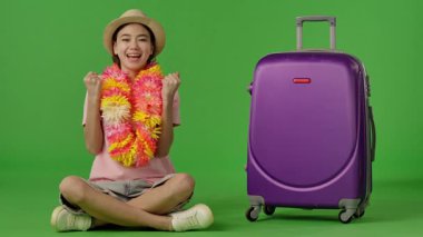 Çiçek kolyeli genç bir kız mor bavulun yanında oturuyor, kameraya bakıyor ve gülümsüyor. Krom anahtar yeşil ekran yeşil ekran stüdyo arka plan modelinde izole edilmiş. Yavaş çekim