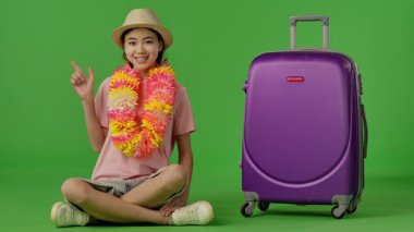 Çiçek kolyeli genç bir kız mor bavulun yanında oturuyor ve parmağıyla onu işaret ediyor. Krom anahtar yeşil ekran yeşil ekran stüdyo arka plan modelinde izole edilmiş. Yavaş çekim