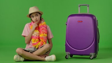 Çiçekli çiçek kolyeli genç kız mor bavulun yanında oturuyor, bir şeyler düşünüyor ve jest yapıyor. Krom anahtar yeşil ekran yeşil ekran stüdyo arka plan modelinde izole edilmiş. Yavaş çekim